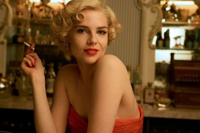 THE RUTH ELLIS STORY An image of Lucy Boynton as Ruth Ellis in A Cruel Love. She is smoking a cigarette, wearing a pink dress and smiling.
