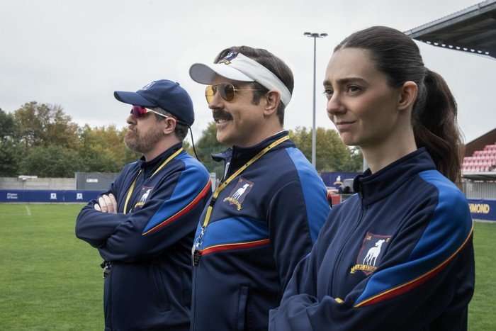 Brendan Hunt, Jason Sudeikis and Tanya Reynolds star in Ted Lasso season 4; their characters are stood in a row on the edge of a football field, looking out at the players
