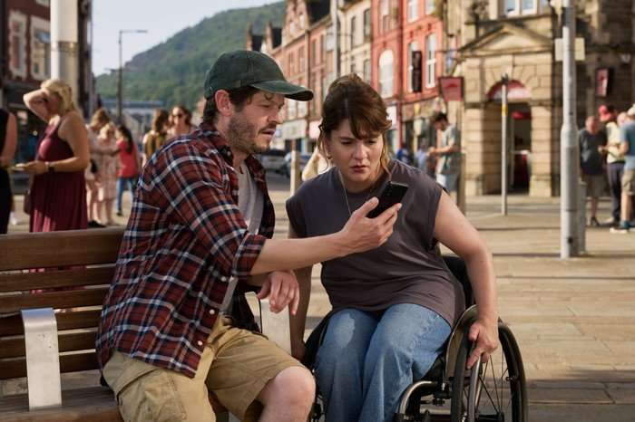 570489,TITLE:The Rapture Iwan Rheon as Carwyn Ruth Madeley as Dr Gabriele Fox in The Rapture looking at a phone screen together whilst sat in the middle of a town.
