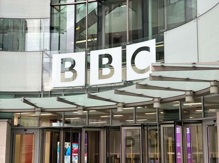 BBC sign on building,Broadcasting House. Exterior of the BBC’s Broadcasting House, showing large white “BBC” logo panels mounted on a glass-and-steel facade above revolving entrance doors.
