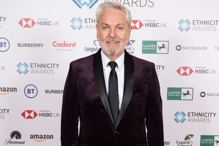 Brian Conley smiling ahead in a black suit and tie.