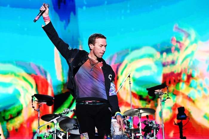 Chris Martin of Coldplay at BBC Radio 1 Big Weekend 2024 Luton Chris Martin of Coldplay performing on stage holding a microphone in the air and dancing, while a colourful display is projected around them.