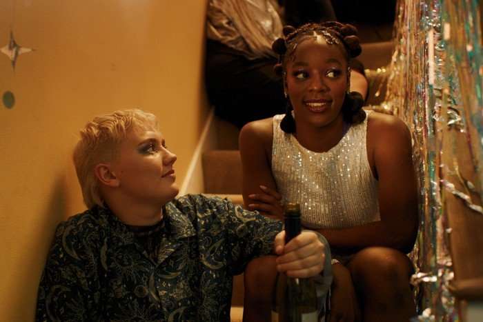 Darcy and Tara sit on a flight of stairs at a house party, wearing glamorous outfits and excitedly talking with friends