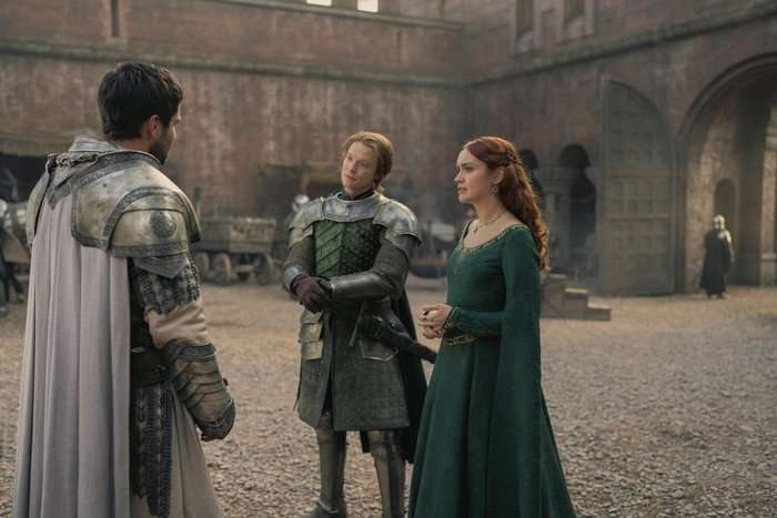 From left to right, Fabien Frankel as Ser Criston Cole, Freddie Fox as Ser Gwayne Hightower, and Olivia Cooke as Dowager Queen Alicent Hightower stand talking in a King's Landing courtyard in House of the Dragon.