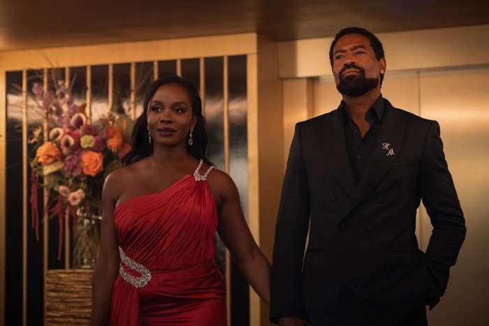 Deborah Ayorinde as Joy & Nicholas Pinnock as Kingsley holding hands whilst walking into a room, both wearing formal attire, Deborah in a red dress and Nicholas in a black suit.