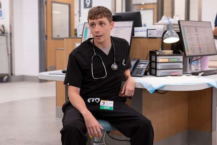 gerran-howell Gerran Howell in The Pitt, wearing hospital scrubs, sat at a desk and with a stethoscope around his neck.