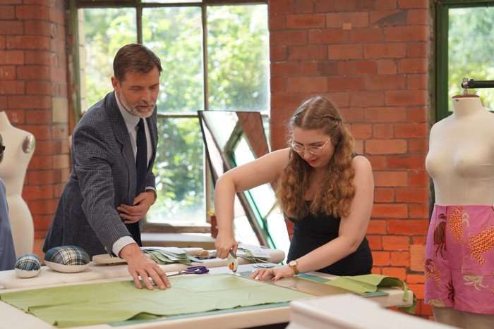 Patrick Grant and contestant Rebecca on The Great British Sewing Bee Patrick Grant is in a grey suit leaning over the work station of Rebecca, who is busy cutting material. He is looking at her work with a hand keeping green material flat as she cuts it