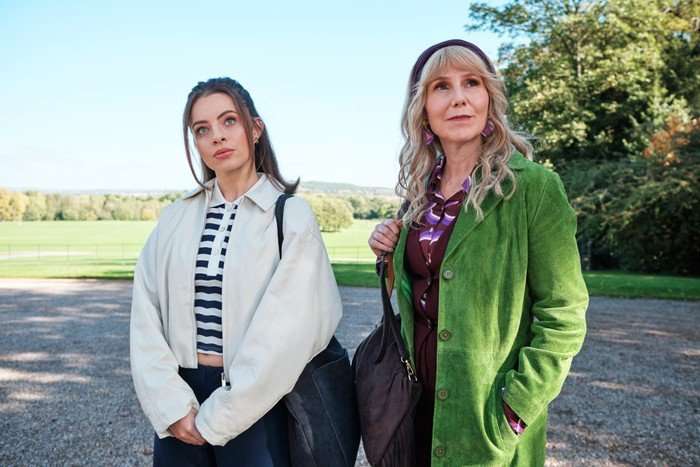 The Hairdresser Mysteries Two people stand outdoors on a gravel driveway with a wide green landscape and trees behind them. On the left, a younger person wears a cream jacket over a navy-and-white striped top and carries a large black shoulder bag. On the right, an older person wears a bright green coat over a patterned purple outfit, a dark headband, and purple earrings, holding a brown shoulder bag. Both are looking off into the distance with thoughtful expressions on a sunny day.