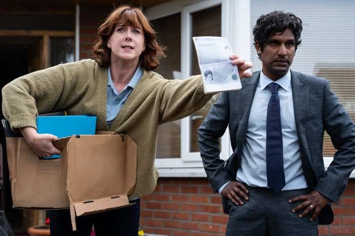 Alexandra Roach (Fran) / Rudi Dharmalingam (Nick) in Hunting Alice Bell. Alexandra Roach (Fran) / Rudi Dharmalingam (Nick) in Hunting Alice Bell standing outside a house, holding a passport and a box of documents, looking angry.