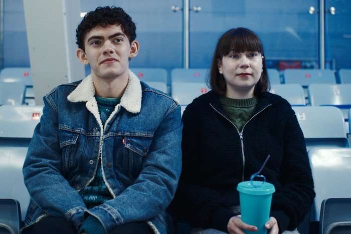 Charlie and Tori sit in the spectator seats at an ice rink, smiling and looking ahead
