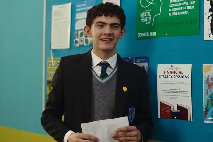 Charlie smiles while wearing his school uniform and standing next to a noticeboard in the school corridor