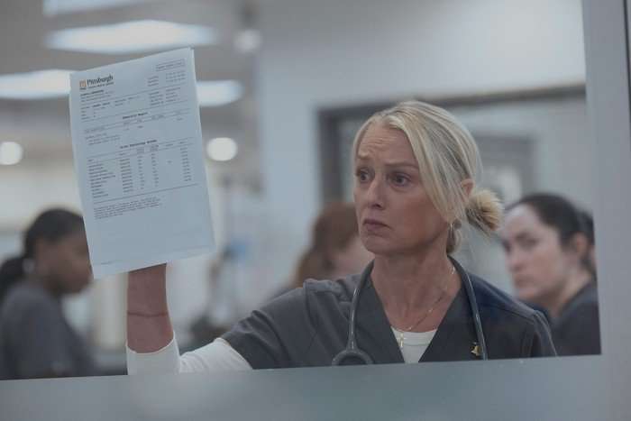 INT. CENTRAL 14 / CENTRAL CORRIDOR SC. 38/39 Katherine LaNasa in The Pitt, stood in a hospital, holding a document up to a glass window.