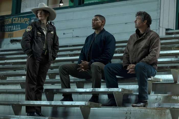 kelly-macdonald-aaron-pierre-kyle-chandler Aaron Pierre and Kyle Chandler sat down on bleachers while Kelly Macdonald stands next to them as they all look outwards.
