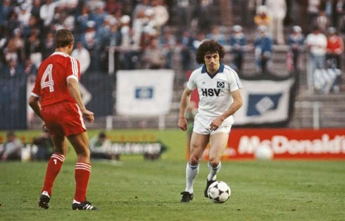Kevin Keegan 1980 European Cup Final Kevin Keegan in a white Hamburg shirt dribbling the ball towards an opponent in red during a match in a packed stadium.