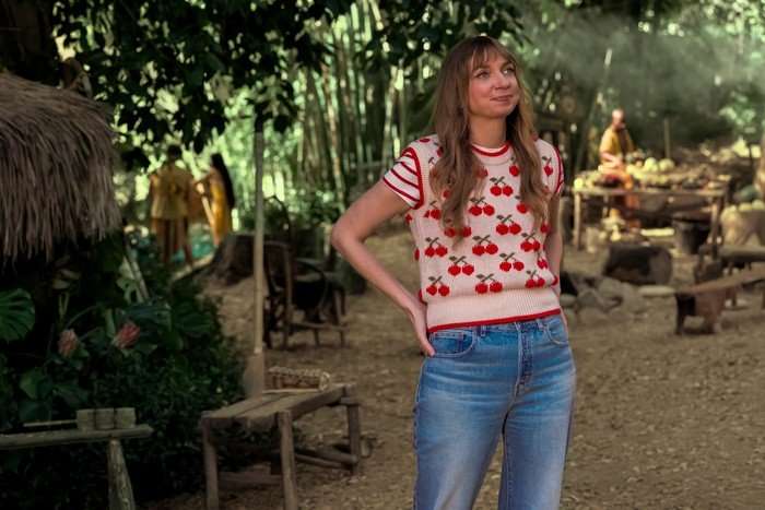 lauren-lapkus-stuart-fails-to-save-universe Lauren Lapkus as Denise standing in the woods, with a fire being lit behind her, wearing blue jeans and a pink jumper vest that has red cherries on it.
