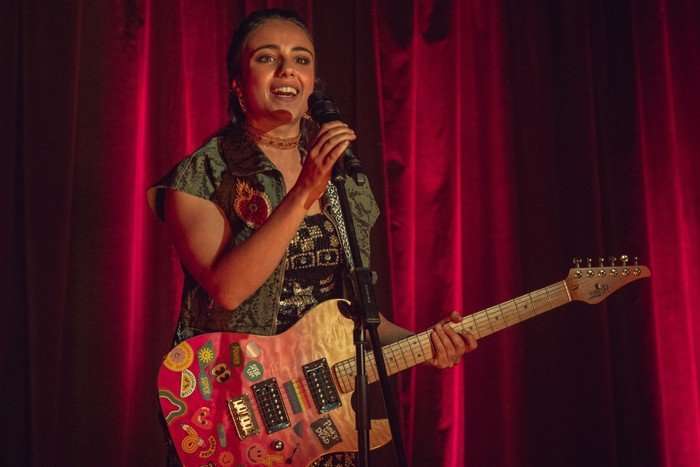 Sahar holds an electric guitar and sings into a microphone on stage, with a red curtain behind her