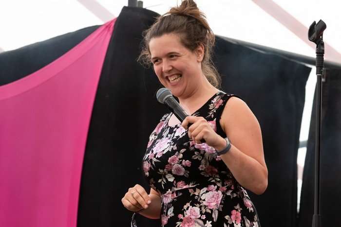 Latitude Festival 2019 Rosie Jones smiles while holding a microphone on stage, wearing a black floral dress against pink-and-black festival draping.