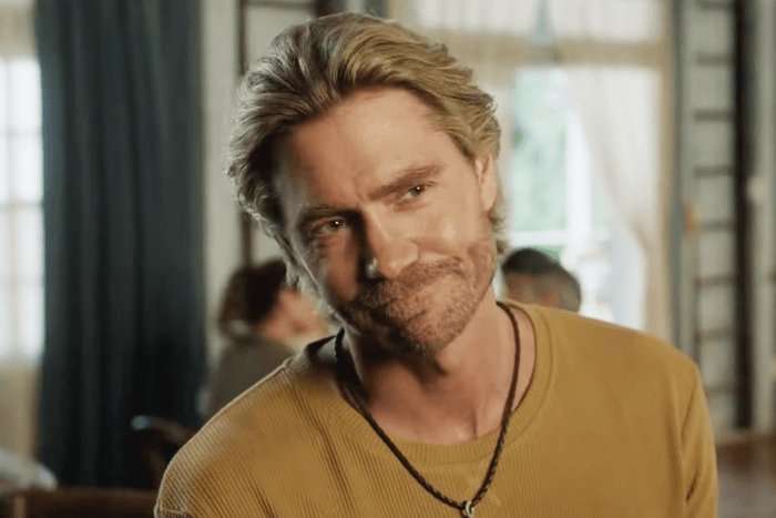 Sullivan's Crossing A close-up of a person with short, swept-back blond hair and light facial stubble smiling warmly with a slight tilt of the head. They are wearing a mustard-yellow ribbed long-sleeve shirt and a black cord necklace with a small pendant. The background is softly blurred, showing what appears to be a cozy café or restaurant with large windows, curtains, and a few seated people, creating a relaxed indoor atmosphere.