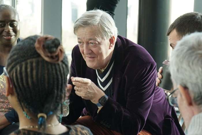 Sir Stephen Fry sat down in a chair leant forward as he points his finger.