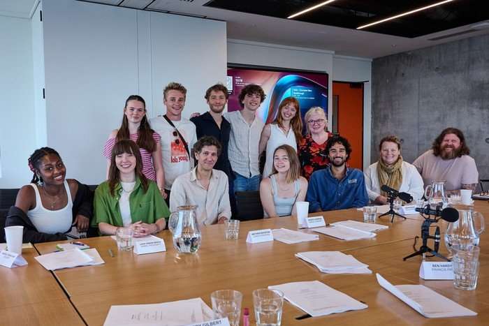 The entire cast of The Cadburys pose together in front of a brown table littered with scripts. Five are sitting down, while six are standing behind them, and they're all smiling