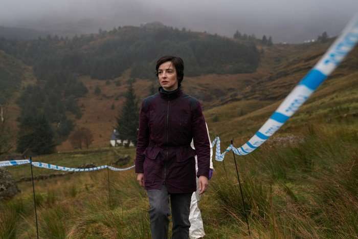 A person wearing a dark purple waterproof jacket walks through a misty, grassy hillside landscape bordered by blue-and-white police tape. Rolling hills and dense pine trees fade into low cloud in the background, creating a bleak, overcast atmosphere. Another figure in a white forensic suit is partially visible behind them, suggesting an active crime scene investigation.