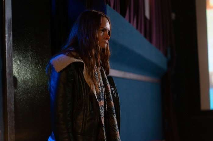 A person with long auburn hair stands inside a dimly lit venue, wearing a black shearling-lined jacket and a multicoloured knitted scarf. They look off to one side with a tense, thoughtful expression, while dark curtains and the edge of a projection screen in the background suggest a cinema, theatre or community hall.
