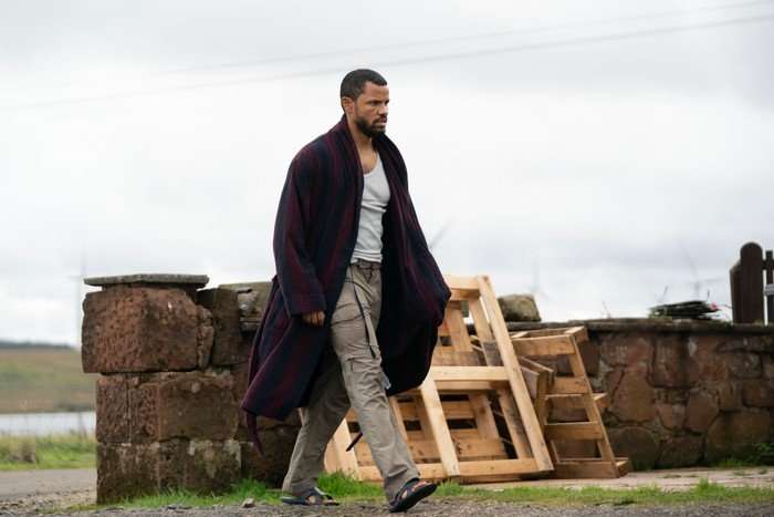 A person wearing a grey vest, beige cargo trousers and sandals walks outdoors wrapped in a dark striped dressing gown. They pass a low stone wall with stacked wooden pallets beside it, under an overcast sky, with open countryside visible in the background. Their serious expression and casual clothing create a sense of urgency or unease.