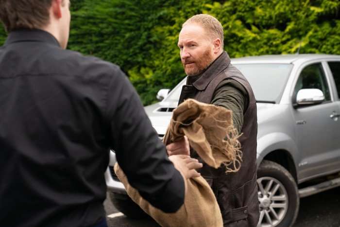 A red-bearded person wearing a dark jacket and brown work vest hands a folded hessian sack to another person whose back is to the camera. They stand in a car park beside a silver pickup truck, with tall green hedges in the background, suggesting a practical exchange taking place outdoors.
