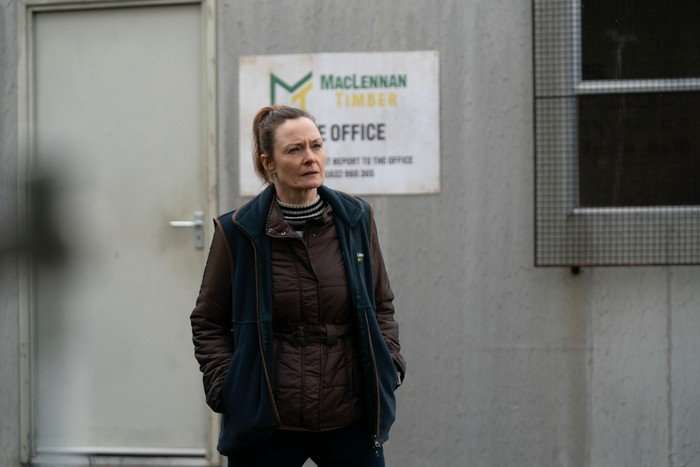 A person wearing a brown quilted jacket, striped high-neck top and dark sleeveless fleece stands outside a portable site office with their hands in their pockets. A sign for "MacLennan Timber" is mounted on the wall behind them, and their focused expression suggests they are watching or waiting in an industrial worksite setting.