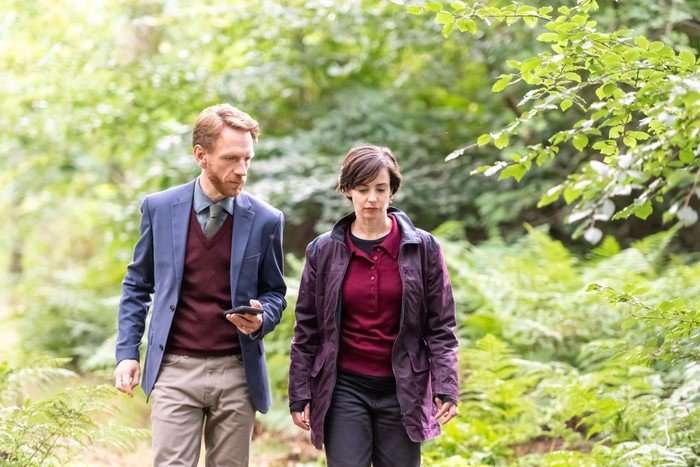 The Dark Two people walk side by side along a woodland path surrounded by lush green trees and ferns. One wears a blue blazer and burgundy jumper while looking at a smartphone, and the other wears a dark purple jacket over a burgundy top, looking thoughtfully ahead. Their serious expressions and quiet conversation suggest they are discussing something important as they move through the forest.