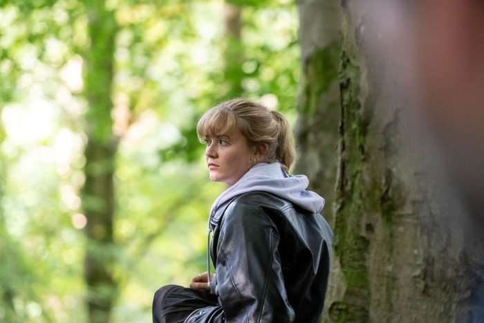 A young person with blonde hair tied back sits against a tree in a sunlit woodland, wearing a black leather jacket over a light grey hooded sweatshirt. They look off into the distance with a pensive, slightly anxious expression, while the softly blurred green forest around them creates a quiet, reflective atmosphere.