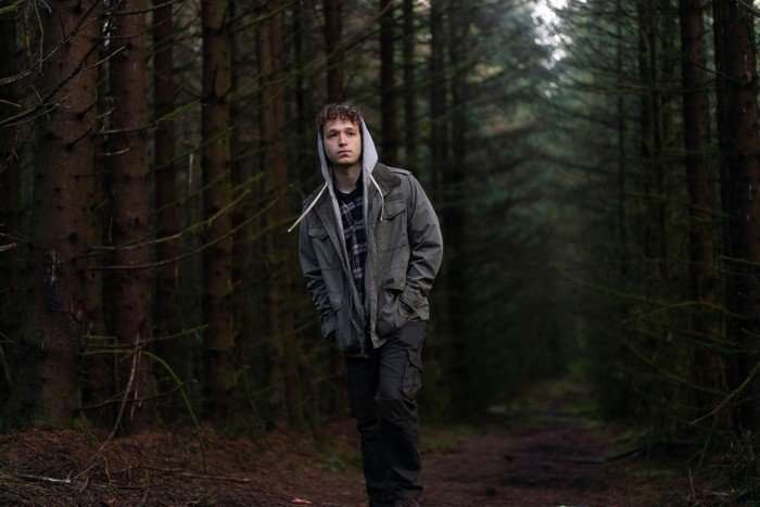 A young person with curly hair walks alone along a narrow path through a dense conifer forest, wearing a grey jacket layered over a hooded sweatshirt and checked shirt. With their hands in their pockets and a serious expression, they move beneath the tall trees, while the dim light and shadowy woodland create a quiet, isolated atmosphere.