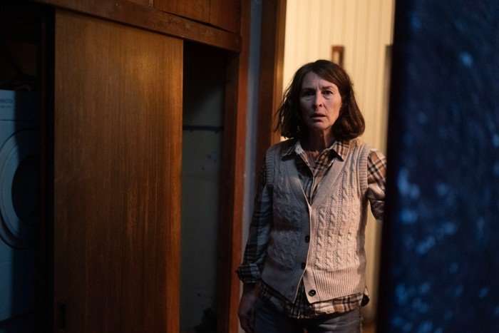 A woman with shoulder-length dark hair stands in a doorway, looking ahead with a tense, startled expression. She wears a beige cable-knit vest over a brown plaid shirt and jeans. Warm light spills from the room behind her, contrasting with the darker foreground and creating an eerie, suspenseful atmosphere.