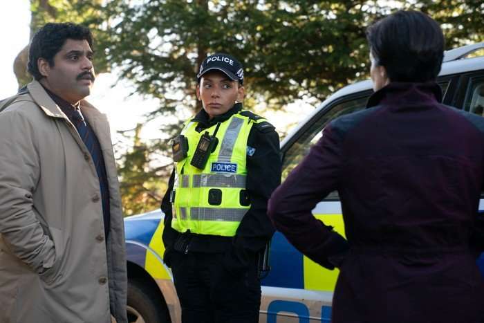 Laura Donnelly as DI Monica Kennedy stands beside a marked police car, speaking with a uniformed police officer and another man in a wooded area in The Dark.