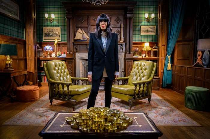 Claudia Winkleman in a tailored black suit stands confidently between two green leather armchairs in a wood-panelled room, facing a table piled high with gold bars.