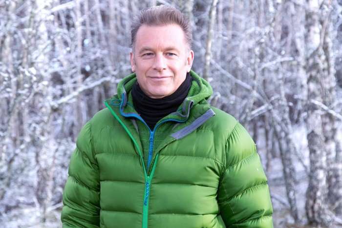 winterwatch (1) Chris Packham stood in a snowy woodland, smiling. He is wearing a green puffer jacket and a black top.